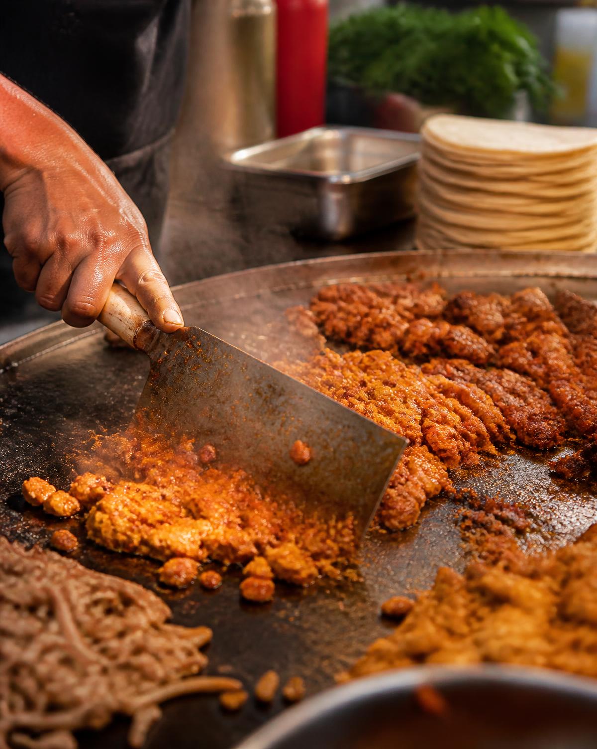 Cook chopping marinated meat on a hot griddle at a taqueria booth